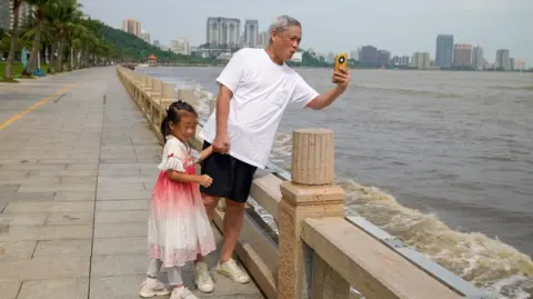 A man in a t-shirt and shorts takes a photo of the ocean while a little girl in a dress holds his hand.