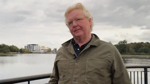 A man with medium length blonde hair, wearing a khaki linen overshirt and a black undershirt, stood against a railing overlooking a body of water in Belfast. Trees and several structures, including the yellow harland and wolff cranes, are visible in the far distance.