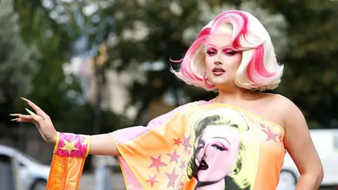 Getty Images Elle Vosque is wearing a blonde wig with pink highlights. She is wearing an orange off the shoulder dress decorated with pink stars and a picture of Marilyn Monroe. 