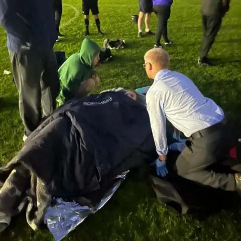 Charlene Paterson Two people kneel around a woman covered in a pile of jackets lying on the grass of a football pitch. It appears to be the evening