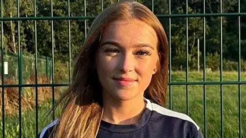 Charlene Paterson A girl with long brown hair smiles at the camera. She stands in front of a wire fence with grass behind it and she appears to be wearing a football strip