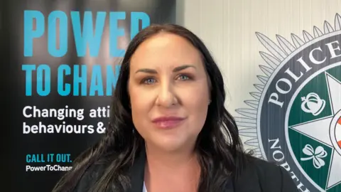 A woman with long brown hair, wearing a black blazer. She is looking at the camera. Half of a PSNI logo is visible on the right hand side of the wall behind her. To the left is a black poster reading "Power to change" in blue. 