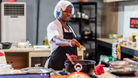 Hilda Baci is standing by a stove and is pinching something into a pot. She is wearing a black apron and has a cloth covering her head.