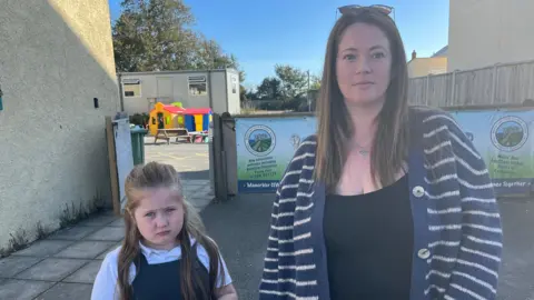 Becky with daughter Isobel. Isobel is wearing a school dress and a white top and has long dark blonde hair, and Becky has long light brown hair and wears a striped cardigan. They are standing next to the temporary school grounds.