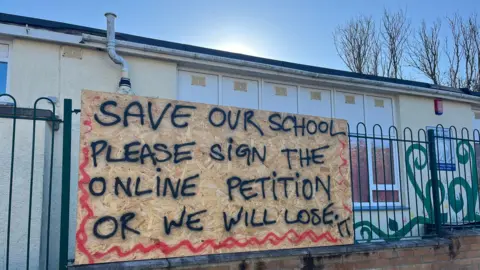 A fence outside the school shows a wooden placard reading: "SAVE OUR SCHOOL PLEASE SIGN THE ONLINE PETITION OR WE WILL LOSE" handwritten in black ink, all in capitals.
