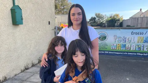 Charlotte and her two daughters Bella and Cleo. They all have long dark hair and the children are wearing school uniform, standing next to their temporary school.