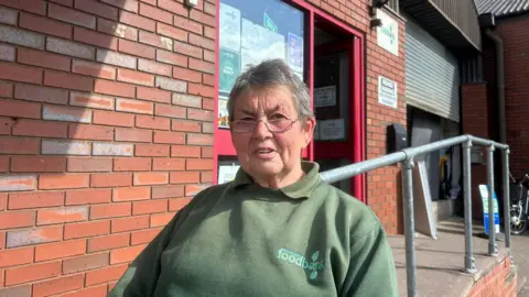 A woman with short grey hair sits outside a red brick building wearing a green sweatshirt. 