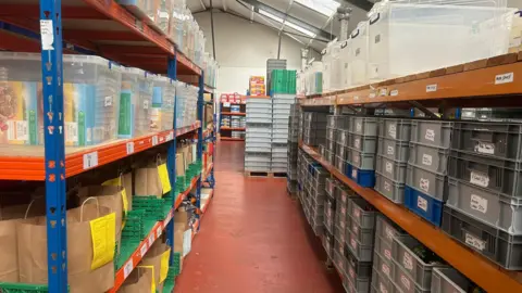 Image taken in the aisle of the warehouse with shelves either side with boxes of cereal in some boxes. 
