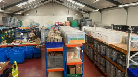 Volunteers sort out food packages next to shelves of food in a large warehouse. 