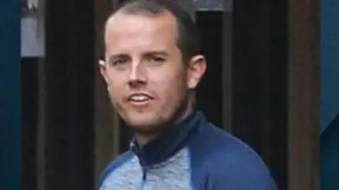 Interpol Head and shoulders image of Sean McGovern. He has short black hair and is wearing a blue and grey tracksuit top. He is smiling and there is an unidentifiable building in the background.