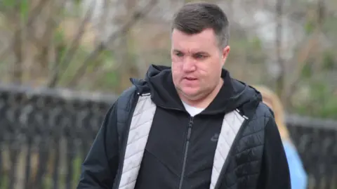 Spindrift Steven "Bonzo" Daniel pictured walking along a street, with a black fence and trees visible but blurred in the background. He is wearing a black hooded jacket and Nike jacket and a white t-shirt. He has short black hair, combed to the side and is looking at the ground. He has extensive facial scarring.