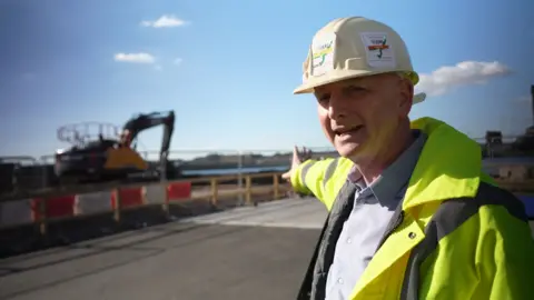 A man in a hard hat and high-visibility jacket stands on a construction site, pointing toward an excavator in the background. Safety barriers line the area, and the sky is partly cloudy.