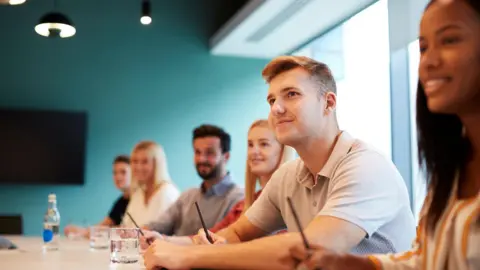 Getty Images A group of people sat at a large meeting table with a pen and paper