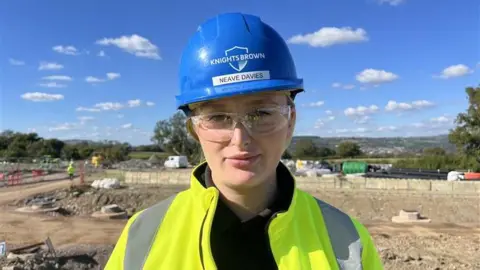 Jack Little on a construction site, wearing a high vis jacket, a blue hard hat, and protective glasses. 