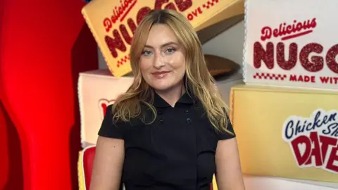 BBC Amelia Dimoldenburg sits in front of some oversized prop chicken nugget boxes, with slogans such as "delicious nuggets" printed on the side. She's wearing a black dress and her blonde hair is down to her shoulders.