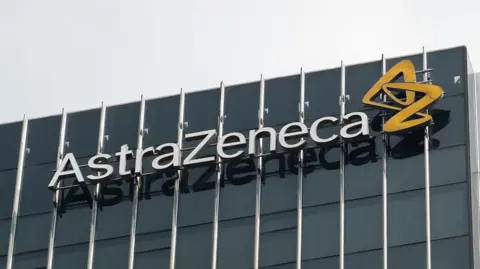 Getty Images Pharmaceutical company Astrazeneca's logo on the side of an office building with dark opaque windows. There is a grey sky behind the building.