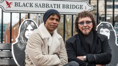 Drew Tommons Carlos Acosta and Tony Iommi sat on Black Sabbath Bridge. The silhouettes of Ozzy Osbourne and Iommi can be seen behind them on the bench. Both of them are looking at the camera, wearing coats and scarves.
