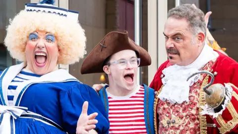Graeme Braidwood Photography A close-up of three panto stars at Royal & Derngate theatre, Northampton. From left Gordon Cooper as the dame in a curly blond wig, wearing bright make-up and a blue and white dress; George Webster as pirate Starky in a brown tricorne hat, red and white stripped top under a blue waistcoat; John Thomson as Captain Hook, with short grey hair, a moustache, wearing an 18th Century-style red jacket over a red gold and white floral waistcoat and holding up his left arm with a hook. 