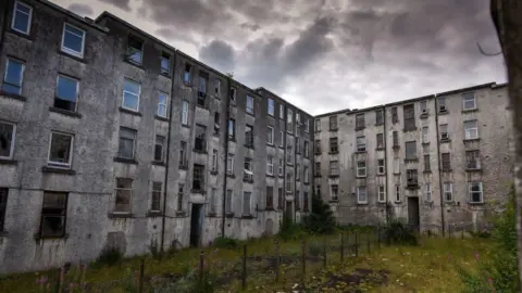 Getty Images Two blocks of abandoned flats in the Clune Park estate in Port Glasgow, pictured under a menacing dark grey sky. The buildings are grey and appear soot stained and overlook a unkempt communal garden.