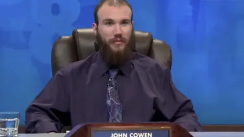 Channel 4 John Cowen has thinning brown hair and a long brown beard. He wears a maroon shirt with purple tie. This is a screengrab from one of his appearances on Channel 4's Countdown programme.