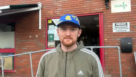 BBC A man wearing a light green tracksuit top and a blue baseball cap stands outside a red brick building. He has a ginger beard. 