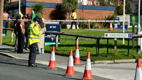 BBC Policewoman and residents stands by orange traffic cones placed outside road entrance into the estate.
