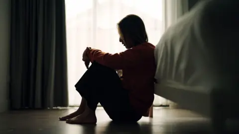 Getty Images The side profile of a woman, sitting on the ground against a bed. There is a window with open curtains behind her. 