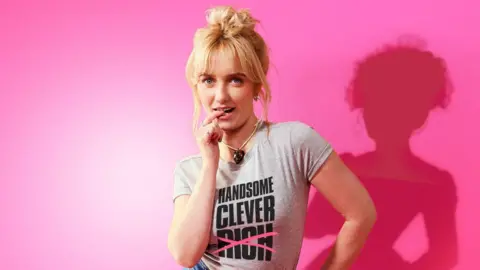 Marc Brenner Amelia Kenworthy as Emma wearing a grey T-shirt with her blonde hair piled up in a bun.