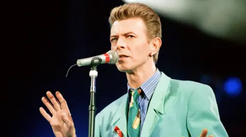 Mirrorpix via Getty Images British Pop Star David Bowie sings into the microphone as he performs on stage while wearing a green suit and tie with slicked back hair against a dark background, at The Freddie Mercury Tribute Concert for Aids Awareness at Wembley in 1992.