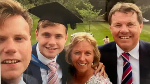 Family handout Jack O'Sullivan, pictured on his graduation day with his family. His brother is on the left of the frame. He has fair short hair and is smiling. Jack is next to him. He also has short fair hair and is wearing a graduation cap and gown over a white shirt and pink and blue striped tie. Catherine is next to him. She has short blonde hair and has glasses on her head. She is looking at the camera and smiling. Alan O'Sullivan is on the right and is wearing a suit.
