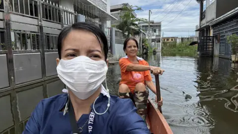 BBC Woman wearing face mask riding a boat through a flooded street with another woman paddling behind her