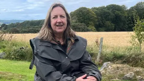 BBC A woman sits in a garden bordered by a field of golden crops. She has shoulder-length blonde/brown hair. She is smiling a the camera and wearing a long green wax jacket. 