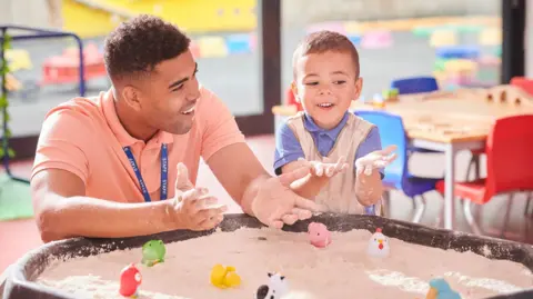 Getty Images A nursery worker wearing a blue lanyard reading "staff" and a small boy wearing an apron play with a sandpit full of plastic animals at nursery. 