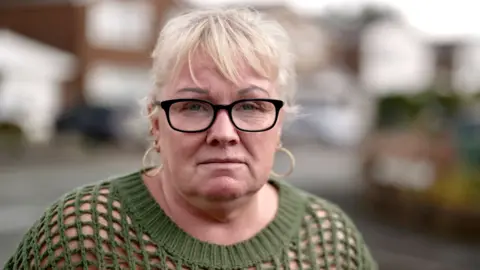 BBC Mandy - a woman with blond hair, gold earrings and black-rimmed glasses. She is wearing a dark-green sweater. She is standing on a street.