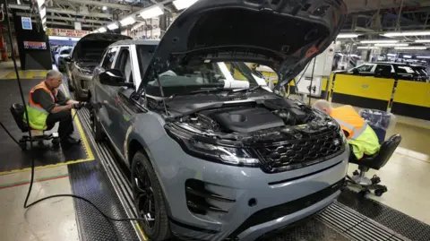 Reuters A grey land rover with its hood open sits on a factory floor, while two workers wearing hi-vis vests work on either side of the vehicle.