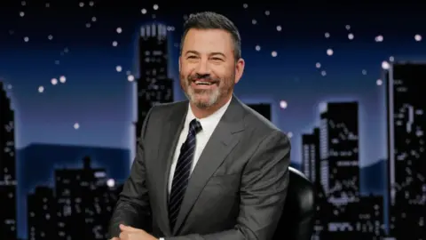 AFP via Getty Images Jimmy Kimmel sits in his seat while presenting his show, smiling and wearing a dark suit and tie.