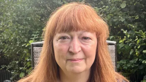 BBC A woman with red hair looks directly at the camera. She is standing in a garden with bushes in the background.