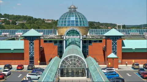 Meadowhall An orange fronted building with glass detail including a large glass dome. There is a sign in white writing which says 'Meadowhall' and surrounding the building is a car park with parked cars. 