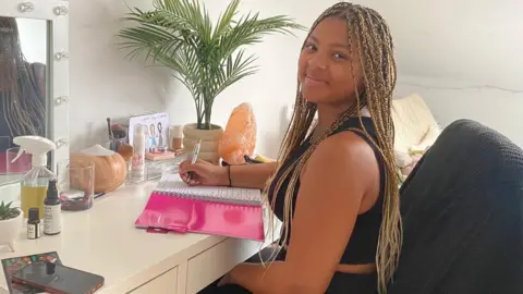 Tianna Hunkins A woman with long braided hair sits at a desk and vanity table in her bedroom, writing notes in her university study book