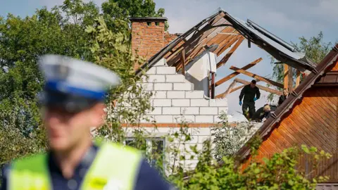 AFP via Getty Images A police officer stands out of focus in the foreground. In the background, people wearing dark clothes and blue latex gloves walk in a hole in a damaged white house, the wooden roof beams exposed.
