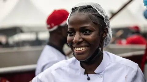 AFP via Getty Images A head and shoulders image of Hilda Baci, wearing a white chef's uniform and hair covering. She has a broad smile on her face.