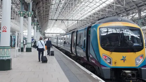 Getty Images Commuters walk along a train platform at Manchester Piccadilly station. A train is currently parked at the platform waiting to pasesengers to board.