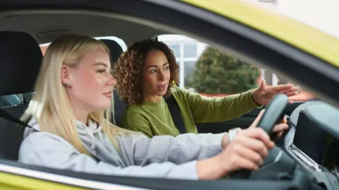 Getty Images Two women in a car. The younger one is at the wheel. She is white and has blonde hair and is wearing a hoodie. An older mixed-race woman is instructing her. The car is yellow-green