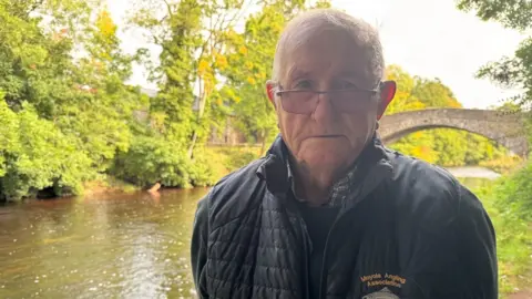 BBC A man with short grey hair wearing glasses and a black jacket that says 'Moyola Angling Association'. He is stood beside a murky river with a stone footbridge and green trees in the background.