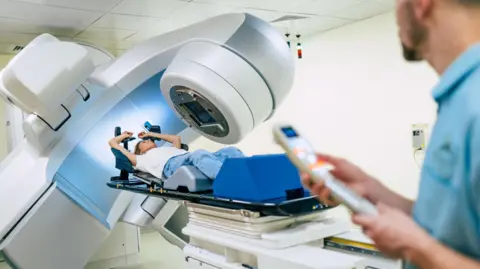 Getty Images A stock image of a woman in a private clinic being giving radiotherapy for cancer. She is lying on her back wearing a white T-shirt and blue trousers with the radiotherapy machine treating her. A radiographer stands to the right of the frame operating the machine with a remote control.