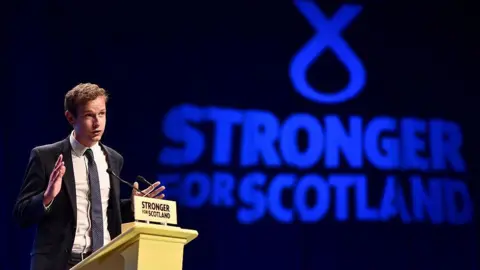 Getty Images Callum McCaig wearing a dark suit over a white shirt and dark tie. He is standing behind a podium. The words 'Strong for Scotland' are in large blue letters behind him.