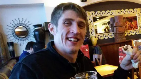 Marshall Family Alan Marshall smiling at the camera. He has dark hair. He is standing and is holding a pint glass. He is wearing a dark jacket.