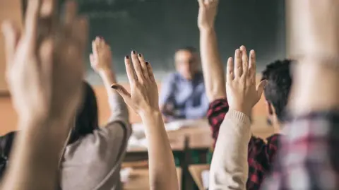 Getty Images Students in a class raise their hands in the air to answer a question. A teacher sits at the front of a class and is blurred in the image. 