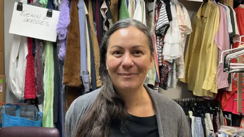 BBC Elk Hargreaves stands in a charity shop, with racks and racks of clothes behind her. She has long dark hair in a ponytail over the shoulder and is smiling.