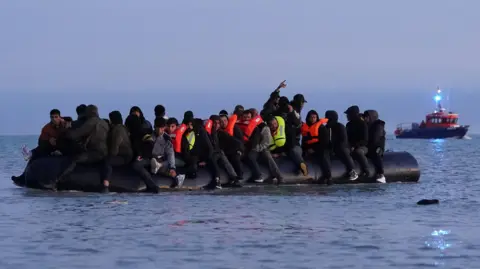 PA Media A black inflatable boat sits low in the water, with people dressed in black and wearing life jackets sitting along the sides of the vessel. In the distance, there is a sturdier red boat with a light shining from its mast. 